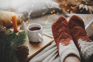 Sıcak çorap giymiş kadın bacakları bir fincan çay, noel yıldızları, altın ışıklar, ağaçlar, mum ve yastıklarla birlikte yumuşak battaniyede. Evde rahat, dinlendirici anlar. Kış ve sonbahar tatilleri.
