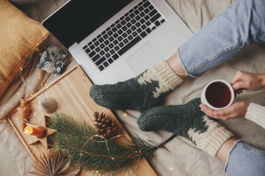 Evdeki sıcak kış tatili. Sıcak bir fincan çayı olan kadın yumuşak yatağında dizüstü bilgisayarla, noel ışıklarıyla, yıldızlarla, çam ağacıyla, mumla ve şık bir odada yastıklarla dinleniyor. Serbest çalışmak ve blog yazmak. Üst görünüm