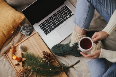 Sıcak bir fincan çayı olan kadın yumuşak yatağında dizüstü bilgisayarla, noel ışıklarıyla, yıldızlarla, çam ağacıyla, mumla ve şık bir odada yastıklarla dinleniyor. Evdeki sıcak kış tatili. Serbest çalışmak ve blog yazmak. Üst görünüm