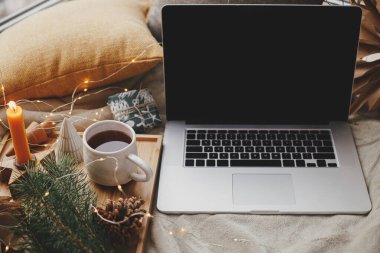 Laptop yumuşak yatakta çay, noel ışıkları, yıldızlar, çam, mum ve İskandinav odasında yastıklarla. Serbest çalışmak ve blog yazmak. Uzaktan kumanda ve çevrimiçi. Evdeki sıcak kış tatili. Boş ekran