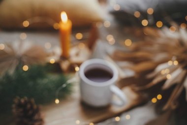 Sıcak bir fincan çayın bulanık görüntüsü. Yılbaşı ışıkları, yıldızlar, çam ağaçları, İskandinav yatak odasındaki mum. Kış kızarıklığı soyut bir arka plan. Rahat bir ev. Atmosferik an