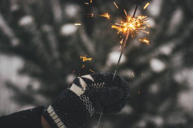 Mutlu yıllar! Karda çam ağacı dallarının arka planında yanan maytap tutan sıcak eldiven. Atmosferik sihir anı. Akşamları elinde parlayan havai fişeklerle kadın eli. İyi tatiller.!