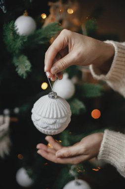 Noel ağacı ışıklarında elleri rahat, modern beyaz süveter tutan eller. Şık Noel ağacını eski süslemelerle süsleyen bir kadın. Kış tatili hazırlıkları. Atmosferik neşeli anlar