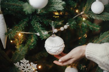 Kış tatili hazırlıkları. Noel ağacı ışıklarında el ele tutuşup, modern beyaz takılar takıyorlar. Şık bir boho noel ağacını klasik süs ile süsleyen bir kadın. Atmosferik hygge ev