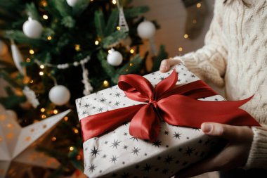 Mutlu Noeller! El örgüsü süveter giymiş eller yılbaşı hediyesini kırmızı kurdeleli ışıklarla sarmış. Şenlikli, süslü İskandinav odasıyla şık bir bayan.