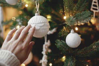 Noel ağacı ışıklarında elinde modern beyaz süveter tutan rahat bir kazak. Şık Noel ağacını eski süslemelerle süsleyen bir kadın. Kış tatili hazırlıkları. Atmosferik neşeli anlar