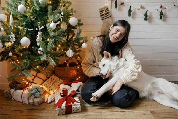 クリスマスツリーの下でギフトやライトとスタイリッシュな幸せな女性が座って巨大な愛らしい犬 お祝いのスカンディナヴィアの部屋で若い女性がかわいい犬を遊んで愛撫 ハッピー ホリデー — ストック写真