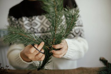 Noel çelengi yapıyorum. Samimi kazaklı bir kadın, çam ağacından bir dal kesiyor. Kırsal arka planda, elleri kapalı. Kış tatili hazırlıkları. Şenlik atölyesi, el yapımı dekor