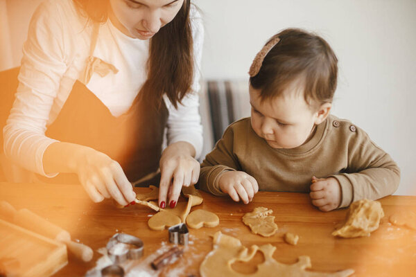 Adorable little daughter with mother making together christmas gingerbread cookies on messy wooden table. Cute toddler girl helps cutting dough with festive cutters for cookies. Family time