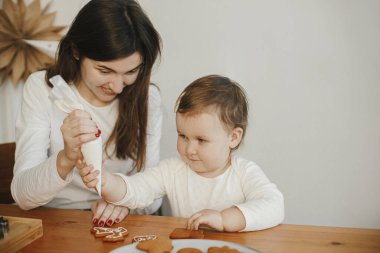 Sevimli küçük kız ve anne birlikte Noel kurabiyelerini modern odadaki ahşap masada kremayla süslüyorlar. Anne kızı otantik güzel anlar. Tatil hazırlıkları