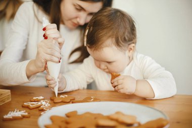 Sevimli küçük kız ve anne birlikte Noel kurabiyelerini modern odadaki ahşap masada kremayla süslüyorlar. Anne kızı otantik güzel anlar. Tatil hazırlıkları