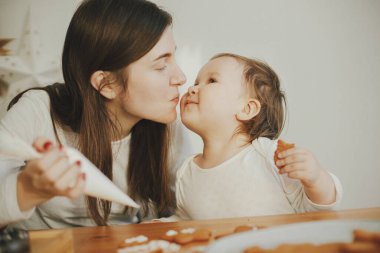 Sevimli küçük kız ve anne birlikte Noel kurabiyelerini modern odadaki ahşap masada kremayla süslüyorlar. Anne kızı otantik güzel anlar. Tatil hazırlıkları