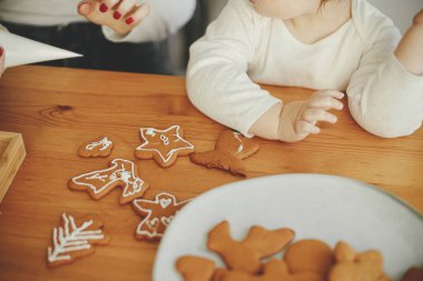 Şirin küçük kız ve anne Noel kurabiyelerini ahşap masa üzerindeki kremayla süslüyorlar, yakın çekim. Aile zamanı, Xmas tatil hazırlıkları. Otantik an