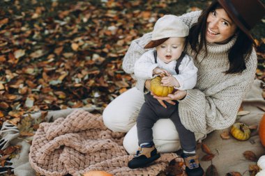 Şık, mutlu bir anne ve sevimli bir oğlan bebek kabak ve sonbahar yapraklarıyla battaniyede oynuyorlar. Sonbahar hasadı ve Şükran Günün kutlu olsun. Sevimli mutlu aile sonbahar bahçesinde dinleniyor.