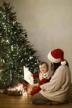 Şirin bebek ve anne rahat süveterler içinde şık bir Noel ağacının altında hediyeler ve şenlikli bir akşamda parlayan ışıklarla oturuyor. Mutlu Noeller ve mutlu tatiller! Aile sihirli xmas arifesinde