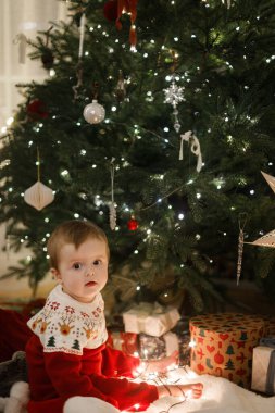 Sihirli Noel arifesinde şirin bir bebek. Mutlu Noeller ve mutlu tatiller! Şen şakrak süveter giyen mutlu çocuk, parlak ışıklarla oynuyor ve süslü Noel ağacında hediyelerle oturuyor.