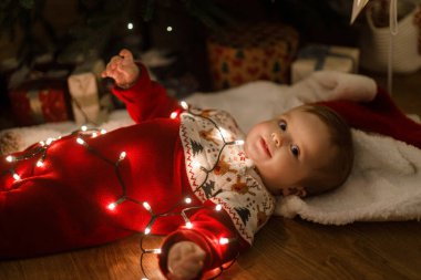 Şık bir Noel ağacının altında parlak çelenklerle süslü süveter giymiş şirin bir bebek. Şenlikli bir akşamda hediyeler, üst manzara. Mutlu Noeller! Mutlu çocuk sihirli xmas arifesinde