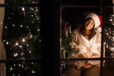 Mutlu Noeller ve mutlu Noeller! Noel Baba şapkalı ve rahat kazaklı bir kadın şık bir Noel ağacında parıldayan yıldızı tutuyor ve akşam penceresinde ışıklar var. Sihirli xmas arifesi dileği