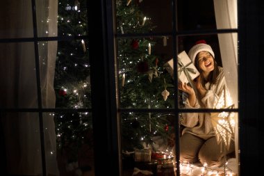 Mutlu Noeller ve mutlu Noeller! Noel Baba şapkalı ve rahat kazaklı bir kadın şık bir Noel ağacında parlak bir hediye kutusu tutuyor ve akşam penceresinde ışıklar var. Sihirli xmas arifesi