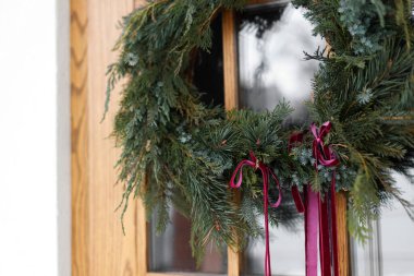 Ahşap ön kapıda kırmızı kurdele asılı şık bir Noel çelengi. Modern çiftlik evinin dış yüzeyinin kış tatili dekorasyonu, fotokopi alanı. Mutlu Noeller.