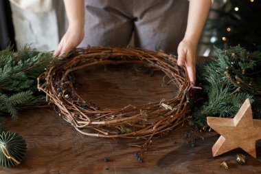 Noel çelengi yapıyorum. Eller çelengi köknar ve sedir dallarıyla düzenliyor, ve makas, kırsal yıldız ahşap masa üzerinde şık xmas ağacının arka planında. Kış tatili hazırlığı ve dekorasyonu