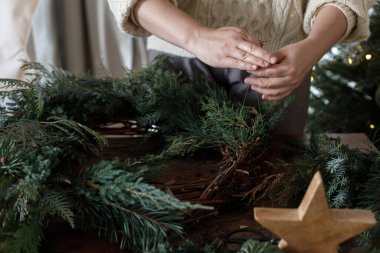 Noel çelengi yapıyorum. Eller çelengi köknar ve sedir dallarıyla düzenliyor, ve makas, kırsal yıldız ahşap masa üzerinde şık xmas ağacının arka planında. Kış tatili hazırlığı ve dekorasyonu