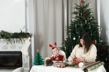 İyi tatiller! Neşeli anne ve şirin bebek Noel hediyelerini oturma odasında Noel ağacı ile paketliyor. Mutlu Noeller! Mutlu aile süveterleriyle şık hediyeler paketliyor.