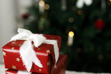Mutlu Noeller ve mutlu tatiller! Süslü, altın ışıklarla süslenmiş Noel ağacına karşı beyaz kurdeleyle paketlenmiş kırmızı Noel hediyesi. Tatil afişi, alanı kopyala
