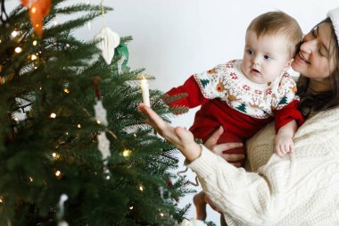 Şen süveter içinde mutlu bir aile modern süslemelerle şık bir Noel ağacı süsler. Mutlu Noeller! Noel Baba şapkalı anne süslü ağaçta şirin bir bebek tutuyor, bayram gelenekleri.