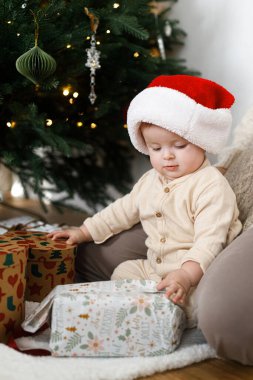 Şirin bebek dekore edilmiş şenlik ağacının altında şık Noel hediyeleri açıyor. Mutlu Noeller ve mutlu tatiller! Pijamalı ve Noel Baba şapkalı mutlu çocuk Noel Baba hediyeleriyle oynuyor.