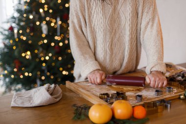 Zencefilli Noel kurabiyesi yapan kadın. Elleri zencefilli ekmek hamuru, oklavalı iğne ve metal kesiciler rustik masa üzerinde Noel ağacının arka planında. Kış tatili hazırlığı
