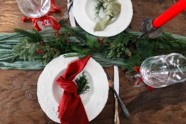 Mutlu Noeller! Noel aile yemeği için şık bir masa. Peçete, cam kaplama, yeşil ve kırmızı kurdeleli modern tabak ve çatal bıçak, köknar dalları ve kırsal masa üzerinde mum.