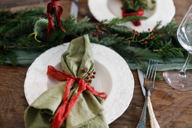 Mutlu Noeller! Noel aile yemeği için şık bir masa. Peçete, cam kaplama, yeşil ve kırmızı kurdeleler, köknar dalları ve kırsal masada mum bulunan modern tabak ve çatal bıçak takımı.