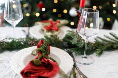 Modern tabaklar, kırmızı ve yeşil peçeteler, çatal bıçak takımı, cam, köknar dalları ve arka plandaki yılbaşı ağacının meyveleriyle süslü bir Noel masası. Mutlu Noeller ve mutlu tatiller!