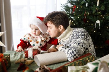 Mutlu bir baba ve Noel Baba şapkalı şirin bir oğlan Noel hediyelerini şık dekore edilmiş ağacın arka planında paketliyor. Mutlu Noeller ve mutlu tatiller! Atmosferik aile zamanı.