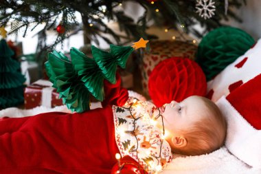 Mutlu Noeller ve mutlu tatiller! Noel ağacının altında hediyelerle kağıt ağacı tutan şenlikli şenlikli şamata içinde mutlu şirin bir bebek ve altın ışıklar. Bebek ilk Noel kutlaması konsepti