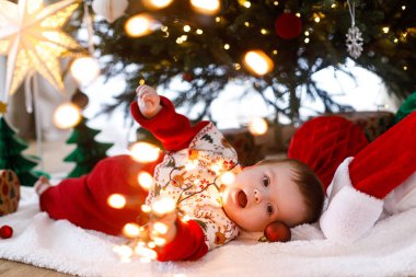 Şenlikli süveter içinde Noel ağacının altında battaniyeye uzanmış altın ışıklarla oynayan tatlı bir bebek. Mutlu Noeller! Atmosferik tatil zamanı, aile süsleme ağacı.