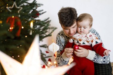 Mutlu Noeller! Mutlu bir baba ve sevimli bir erkek bebek süveter giyip noel ağacını modern takılarla süslüyorlar. Neşeli aile zamanları, kış tatili hazırlıkları