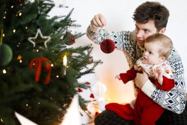 Mutlu baba kucağında tatlı bir çocuk ve süslü Noel ağacını kırmızı mücevherle süslüyor. Mutlu Noeller ve mutlu tatiller! Atmosferik aile zamanı, tatil hazırlığı.