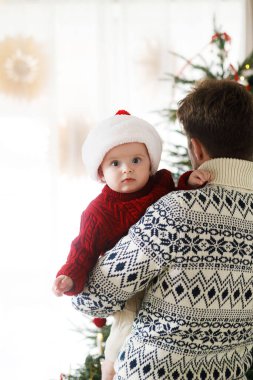 Babam Noel Baba şapkalı şirin bebeğe sarılıyor ve Noel ağacı ışıklarının arkasında süveter örüyor. Tatil aile zamanı, tatlı anlar. Mutlu Noeller ve mutlu tatiller!