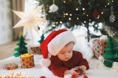 Noel Baba şapkalı ve kırmızı örgü süveterli sevimli bebek altın ışıklarla süslenmiş Noel ağacının altında takılarla oynuyor. Bebeğin ilk Noel 'i, tatlı anları. Mutlu Noeller.!
