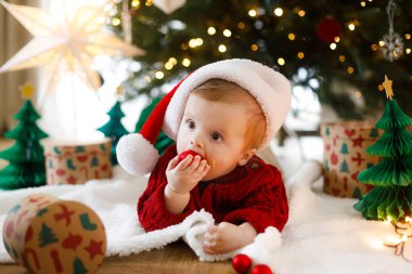 Noel Baba şapkalı ve kırmızı örgü süveterli sevimli bir çocuk, altın ışıklarla süslenmiş Noel ağacının altında takılar yiyor. Bebeğin ilk Noel 'i, tatlı anları. Mutlu Noeller.!
