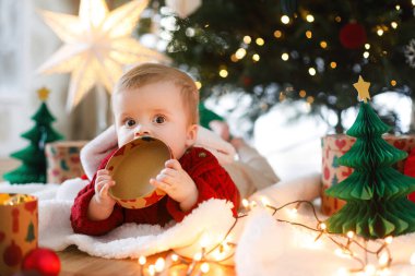 Kırmızı örgü süveter giymiş şirin bir bebek altın ışıklarla ve hediyelerle şık bir Noel ağacında hediye kutusuyla oynuyor. Mutlu Noeller ve mutlu tatiller! Bebek Noel Baba