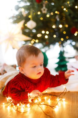 Kırmızı örgü süveter giyen sevimli bebek Noel ağacının altında çelenk ışıklarıyla oynuyor. Bebeğin ilk Noel 'i. Mutlu Noeller.!