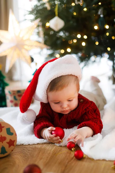Noel Baba şapkalı ve kırmızı örgü süveterli sevimli bebek altın ışıklarla süslenmiş Noel ağacının altında takılarla oynuyor. Bebeğin ilk Noel 'i, tatlı anları. Mutlu Noeller.!