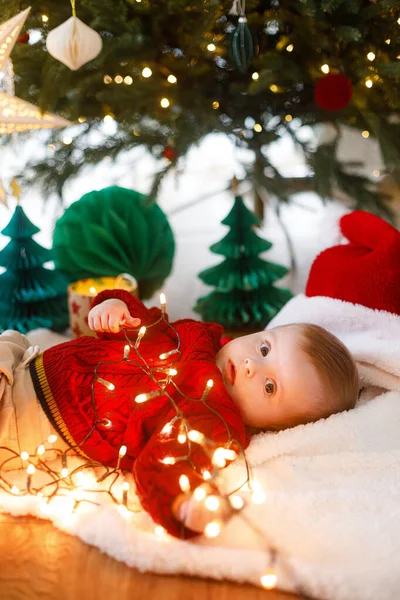 Kırmızı örgü süveter giyen sevimli bebek Noel ağacının altında çelenk ışıklarıyla oynuyor. Bebeğin ilk Noel 'i. Mutlu Noeller.!
