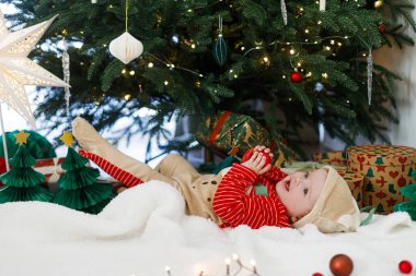 Noel kostümlü şirin bebek kırmızı takılarla oynuyor ve süslü Noel ağacının altında hediyeler ve ışıklarla yatıyor. Mutlu Noeller ve mutlu tatiller! Bebeğin ilk Noel 'i