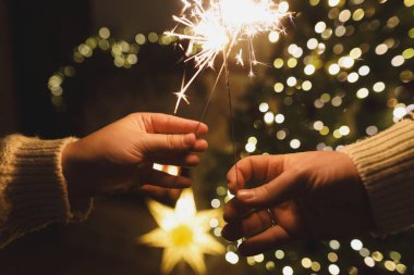 Noel ağacı ışıklarına karşı havai fişek tutan eller akşam sıcacık oturma odasında sallanıyor. Yeni yılınız kutlu olsun! Atmosferik aile kutlaması, ellerinde yanan maytaplarla.