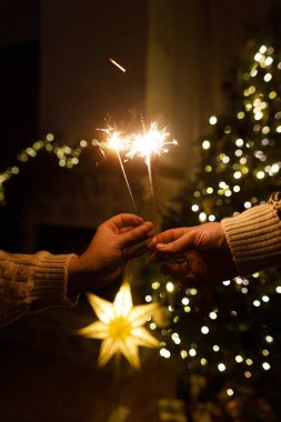Noel ağacı ışıklarına karşı havai fişek tutan eller akşam sıcacık oturma odasında sallanıyor. Yeni yılınız kutlu olsun! Atmosferik aile kutlaması, ellerinde yanan maytaplarla.