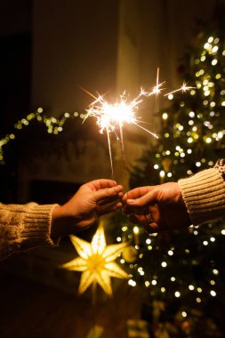 Noel ağacı ışıklarına karşı havai fişek tutan eller akşam sıcacık oturma odasında sallanıyor. Yeni yılınız kutlu olsun! Atmosferik aile kutlaması, ellerinde yanan maytaplarla.
